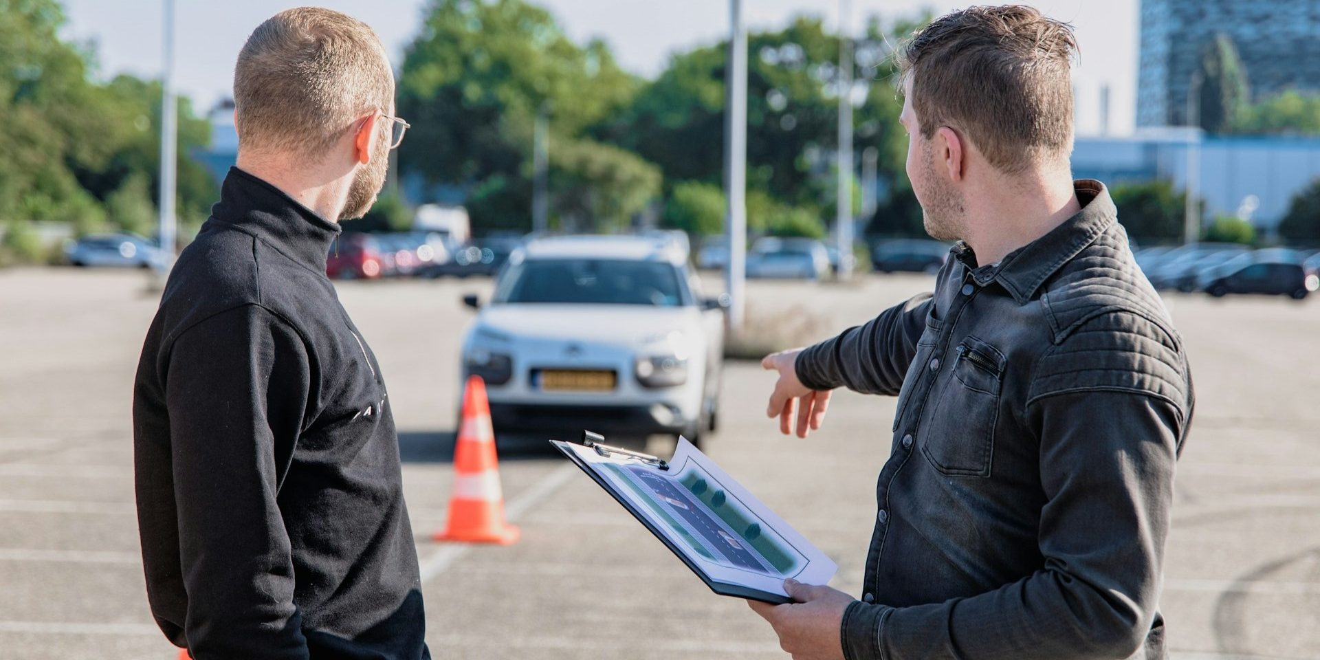 Instructeur wijst naar oefenauto tijdens rijles op parkeerterrein met verkeerskegels – ontspanning na rijles.