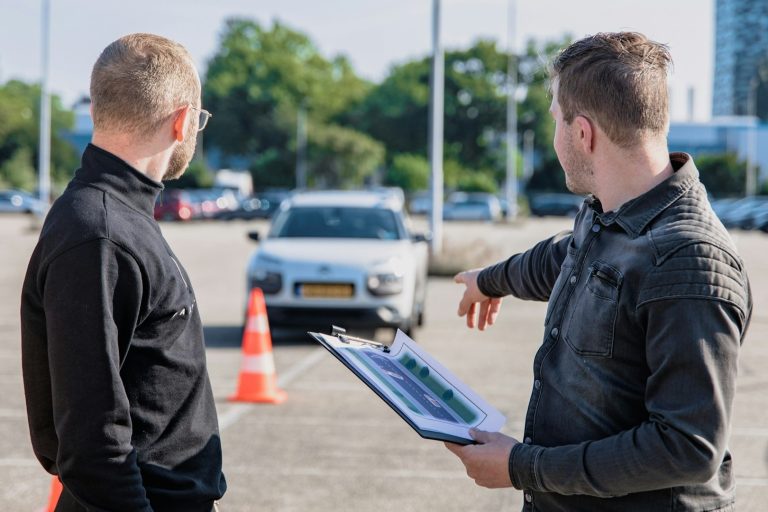 Instructeur wijst naar oefenauto tijdens rijles op parkeerterrein met verkeerskegels – ontspanning na rijles.